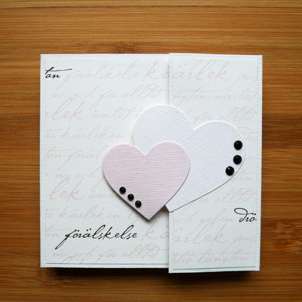 Vitt fotoalbum med mönstrat papper som har snirkliga ord i rosa och svart skrivet över sig. Ett rosa och ett vitt hjärta är placerat vid öppningen av albumet. Tre svarta dekorstenar är limmade på vardera hjärta.