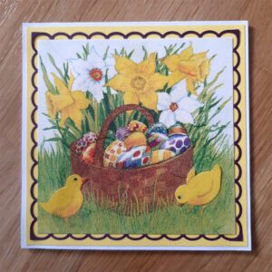 Kvadratiskt kort i färgerna vitt, gult, lila och grönt. Motiv av en korg fylld med ägg som står på en gräsmatta med två kycklingar som pickar runt om.